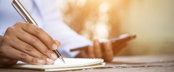 Image of the hands of a black woman writing in a notebook while holding a cell phone