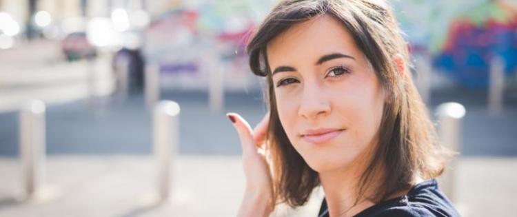 Photo of a young woman looking at the camera, tucking her hair behind her ear.