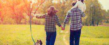 A man and woman holding hands while walking their dog and carrying their daughter in a park