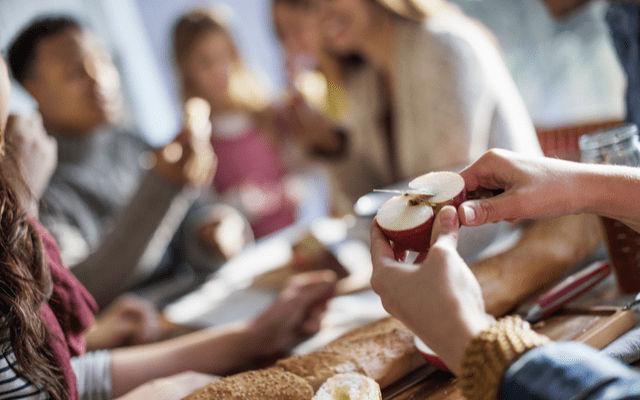 Photo of hands slicing an apple at a food-covered table surrounded by people