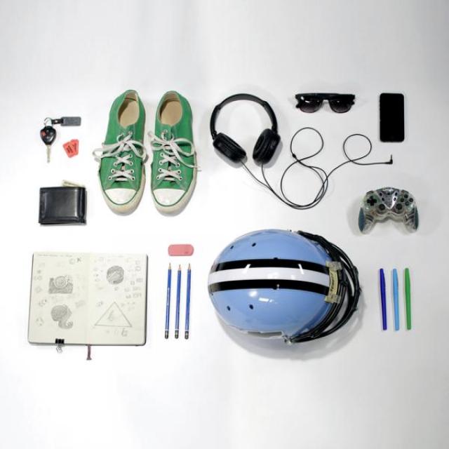 Lots of everyday objects are laid out on a white background. A wallet, car keys, a notebook, shoes, headphones sunglasses, pens and pencils, and even a video game controller are all set down in an orderly fashion. 