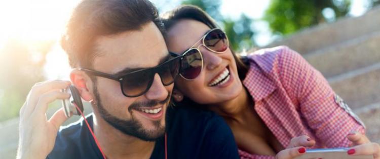 Photo of a man sitting outside on steps listening to headphones. His girlfriend sits next to him, holding her phone.